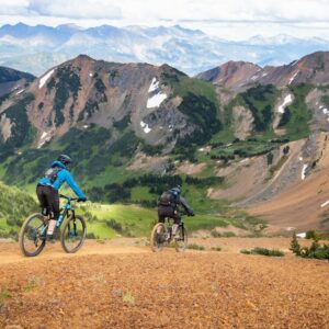 Two mountain bikers riding on a scenic alpine trail with mountains in the background.