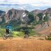 Two mountain bikers riding on a scenic alpine trail with mountains in the background.