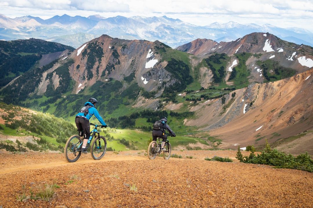 Two mountain bikers riding on a scenic alpine trail with mountains in the background.
